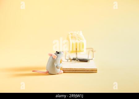 Small plasticine gray mouse looks at a piece of cheese in a mousetrap. Beige background. Stock Photo