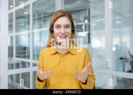 Mature adult business woman with blond hair working inside light office, customer service tech worker smiling and talking to customer consulting, using headset and laptop for remote video call. Stock Photo