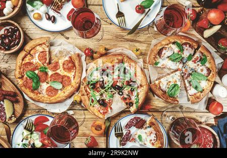 Pizza party table. Top view glasses with red wine, rustic wooden table with hot pizzas, italian appetizers, salads, cheese, fruits and berries. Family Stock Photo