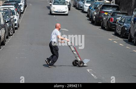 Delivery driver man pushing empty trolley across a road after delivering food to a retailer in England, UK. Stock Photo