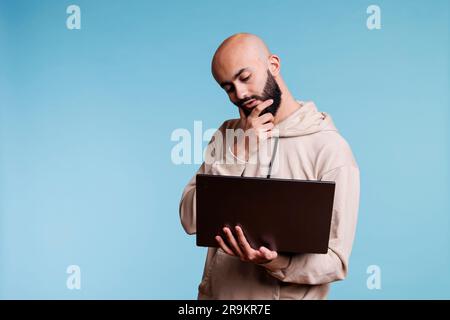 Thoughtful arab man rubbing chin in doubt while working on laptop. Young pensive person with puzzled expression thinking while holding portable computer and analyzing business data Stock Photo