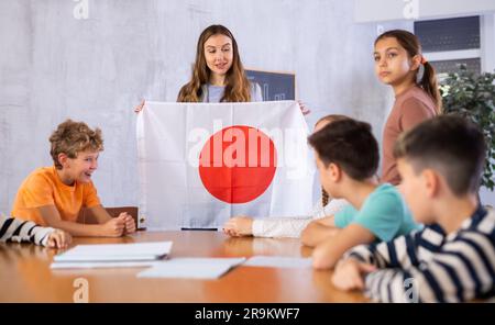 Joyful young woman teacher demonstrating flag for Japan schoolkids preteens during lesson of geography in auditory Stock Photo