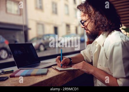 Male student making notes during online lecture while sitting in cafe near window Stock Photo