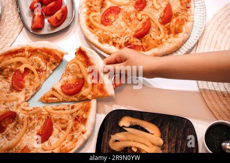 Homemade vegeterian pizza with cheese, tomatoes and pepper on kitchen table. Top view of Little girl hand taking a piece of pizza away to eat. Stock Photo