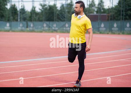 Young man with wireless headphones warming up leg muscles before training on running track Stock Photo