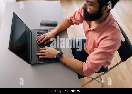 Top view of successful Indian programmer male in eyeglasses working on laptop sitting at desk in light coworking office room. Stock Photo