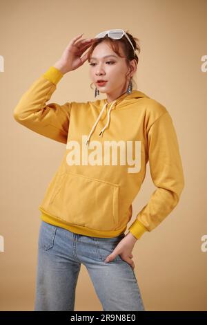 Portrait of transgender young woman in yellow hoodie and jeans posing against beige background Stock Photo
