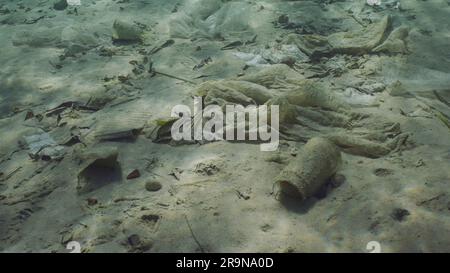 Plastic garbage lies on sandy-silty bottom of sea in shallow water on bright sunny day in sun glare. Seabed polluted with plastic and other debris, Re Stock Photo