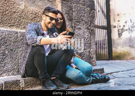 Seated on an urban sidewalk, a young couple shares a laugh while looking at funny content on a smartphone. Stock Photo