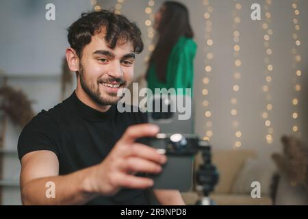 Positive bearded man in black shirt smiling and taking photo on smartphone while sitting in cozy room Stock Photo