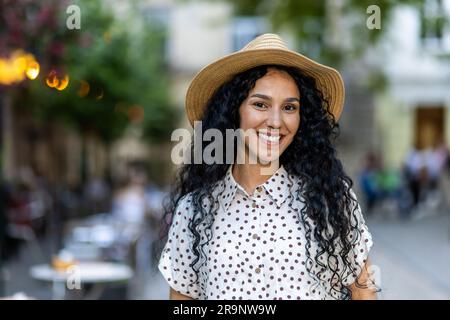 Beautiful young Latin American woman portrait, woman walking in evening city in hat with curly hair in warm weather, smiling and looking at camera close up. Stock Photo