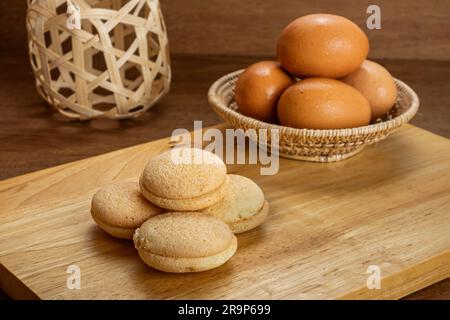 High angle view pile of small homemade sweet cake filled with pineapple jam on wooden board with pile of chicken eggs in bamboo basket on wooden table Stock Photo