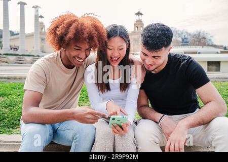 Group of diverse friends using a cellphone to watch and share on the social media funny pics. Three young adult people or teenage students sitting outside having fun with a smartphone messaging app. High quality photo Stock Photo