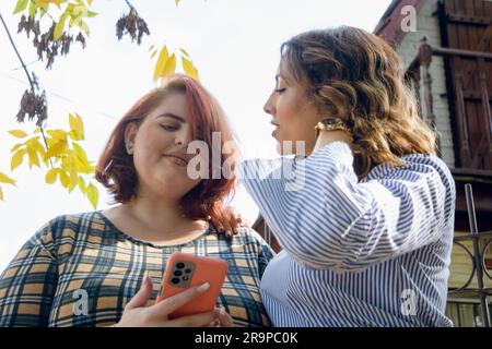 Two young Latina friends of Argentinian ethnicity, business women, standing talking to each other, dressed in casual clothes, using the phone outside Stock Photo