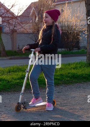Child riding on kickscooter alone. Blonde cute girl on scooter on spring city park street background. Active lifestyle leisure, physical sports traini Stock Photo