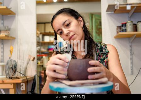 Woman in pottery studio working raw ceramic and clay to make pot. Pottery workshop with natural light scraping, smoothing, shaping and sculpting. maki Stock Photo