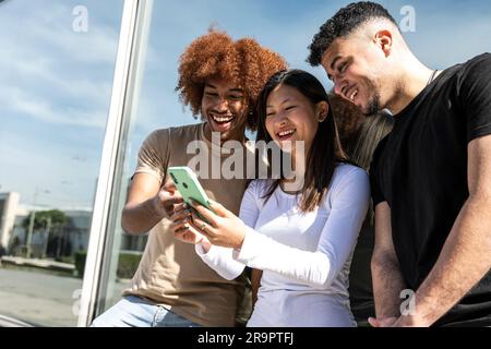 Three young diverse people looking at a smartphone standing int the street.Two young men smiling and watching at the phone of a happy woman. Stock Photo