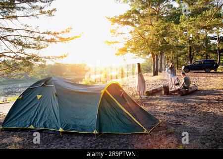 Tourist tent in a picturesque place on the banks of the river against the backdrop of a sunset in summer. A group of friends are resting in the forest Stock Photo