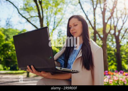 Outdoor portrait of young business woman working in spring park on laptop. Female entrepreneur uses internet in public garden Stock Photo