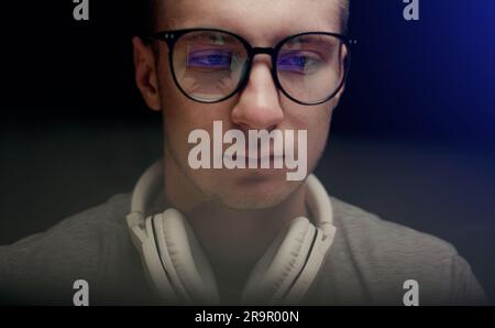Concentrated young professional programmer in computer glasses with monitors reflection works online late. Serious hacker, IT specialist using laptop computer. Esport player in virtual competition. Stock Photo