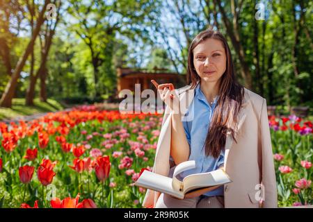 Young caucasian businesswoman smiling holding notebook in spring park pointing. Entrepreneur having break outdoors relaxing in public garden. Space Stock Photo