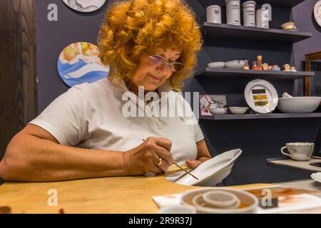 A craftswoman with red curly hair hand-paints a porcelain plate with orange on-glaze paint in the demonstration workshop of the Porcelain Museum of the Meissen Porcelain Foundation, Saxony, Germany. Additional hand-painted porcelain pieces stand on a shelf in the background. Stock Photo