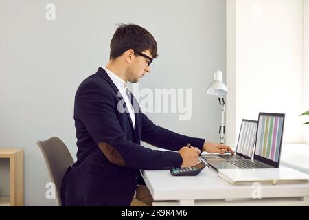Young concentrated male business financier working with spreadsheets using two laptops. Stock Photo
