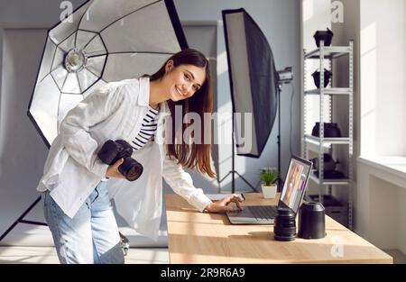 Smiling woman photographer with camera editing photos on laptop in office Stock Photo