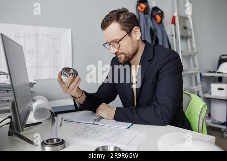 Portrait of bearded mechanical engineer holding machine part at workplace while reviewing blueprints Stock Photo