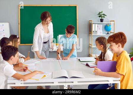Young smiling teacher is engaged with student standing at table against the background of blackboard. Stock Photo