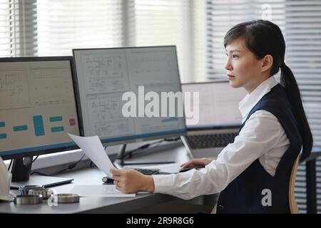 Side view portrait of Asian woman as female engineer using computer with blueprints on screen while doing calculations in office, copy space Stock Photo