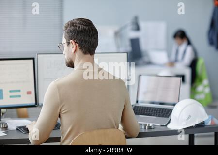 Back view of bearded adult man working as engineer at workplace with hardhat on table, copy space Stock Photo