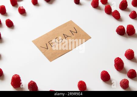 The word vegan is written on paper and lies on a white background and around lies raspberries, spread out on a white background, without meat, fruits Stock Photo