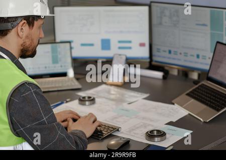 Side view of male engineer wearing hardhat at workplace in office and using computer designing blueprints, copy space Stock Photo