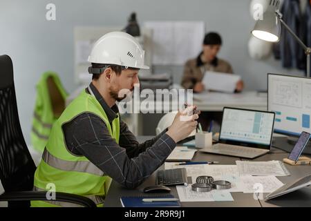 Side view portrait of male engineer holding steel bearing machine part while working on construction plans in office, copy space Stock Photo