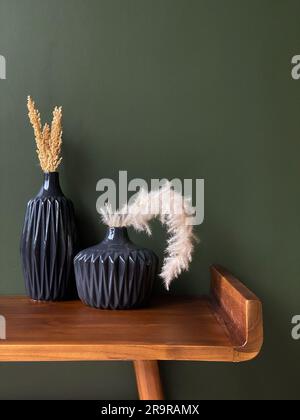 Two black vases with dried grass and plants on a wooden table against a green wall Stock Photo