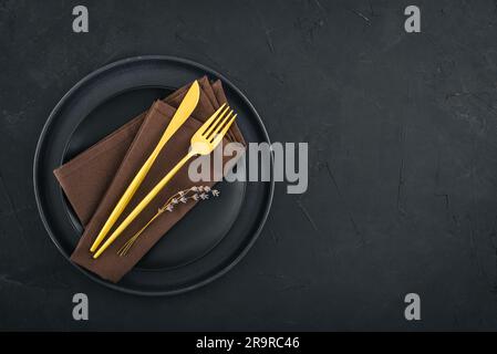Modern minimal table place setting with black plates and golden cutlery on black  background top view. Space for text . Modern kitchen. Stock Photo