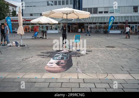 Kaiserslautern, Germany. 28th June, 2023. Rafael Reyes standing with smartphone in front of his impressive painting. Eleven international artists from 6 countries are turning the Stiftsplatz (Square) pavement into three-dimensional art. 3D street art uses perspective to create optical illusion of space. Visitors are welcome to watch all artists at work over three days from Wednesday at 8:00 AM to Friday 6:00 PM. This years topic is 'Digitalisation and Smart City'. Paintings will depict robots, data, or cyberpunk scenarios. Credit: Gustav Zygmund/Alamy News Stock Photo