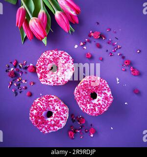 Flatlay of raspberry doughnuts on square purple background with fresh pink tulip bouquet and dries rose petals stock photo Stock Photo