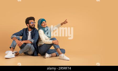 Nice Offer. Smiling Muslim Couple Sitting On Floor And Pointing Aside Stock Photo