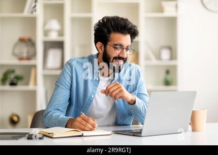 Online Education. Young Indian Man Watching Webinar On Laptop And Taking Notes Stock Photo