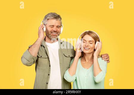 Happy european middle aged couple listening to music with closed eyes, man hugging wife, posing over yellow background Stock Photo