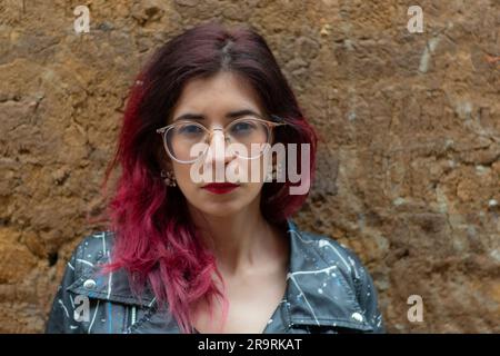 A young woman poses in a black leather jacket, her bright red dyed hair a striking contrast against the grey concrete wall behind her Stock Photo