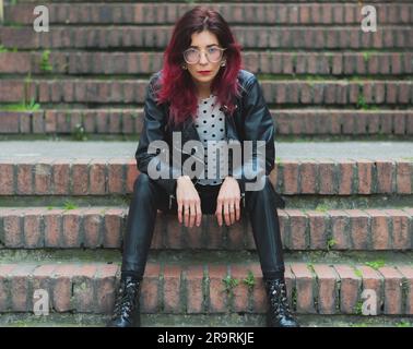 A young, attractive woman is featured wearing a stylish leather jacket and black boots while seated on a staircase Stock Photo