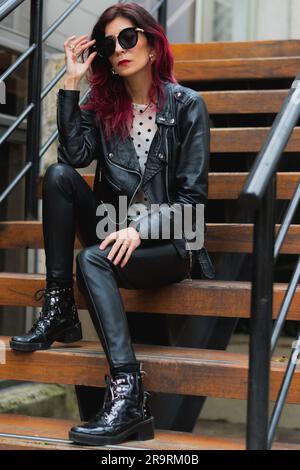 A young woman sits on the steps of a staircase, dressed in black trousers and boots Stock Photo