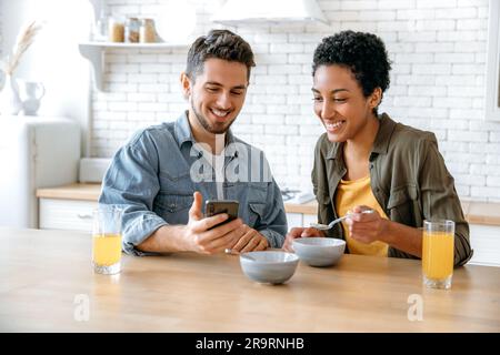 Using gadgets during breakfast. Happy couple in love, african american woman and caucasian man, using mobile phone during morning breakfast at home, watching online videos or news, smile Stock Photo