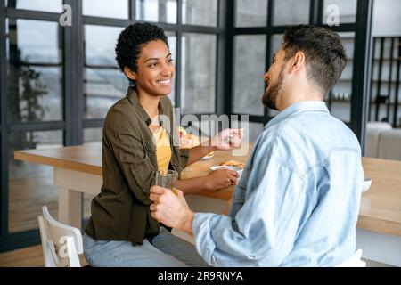 Spending time together. Happy family couple, caucasian man and african american woman, having breakfast at kitchen with healthy porridge, spend time together at weekend morning, communicating, smile Stock Photo