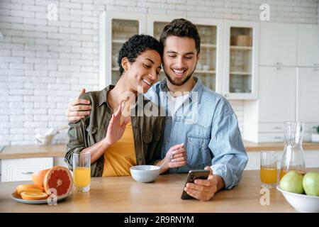 Online video call. Cheerful loving couple of different nationalities, talking via video conference using smart phone, during a morning breakfast, looking into the camera of the mobile phone, smiling Stock Photo