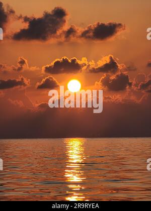 A stunning sunset scene featuring a blazing orange sun setting behind a sky filled with clouds and a tranquil body of water in the foreground Stock Photo