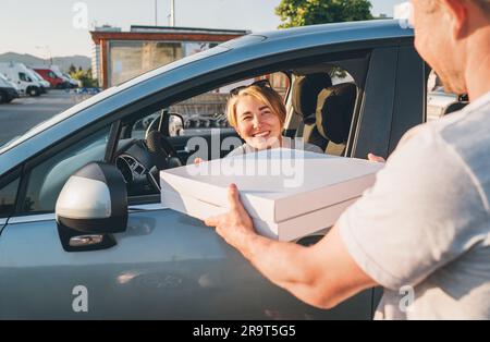 Young female sitting in the car driver seat and smiling to deliveryman accepting two pizza boxes on the city street. Small business, people relations, Stock Photo